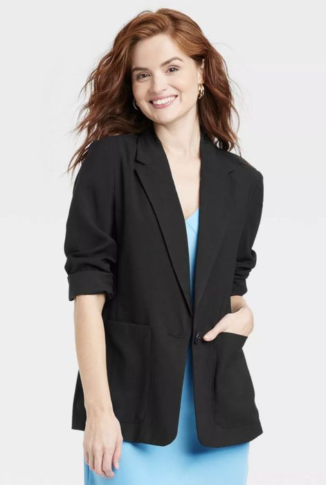 A person smiling, wearing a black blazer over a blue dress, with hands casually tucked in the pockets of the blazer