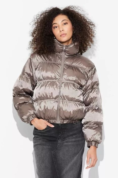 A person standing against a plain background, wearing a puffer jacket and jeans, with a neutral expression