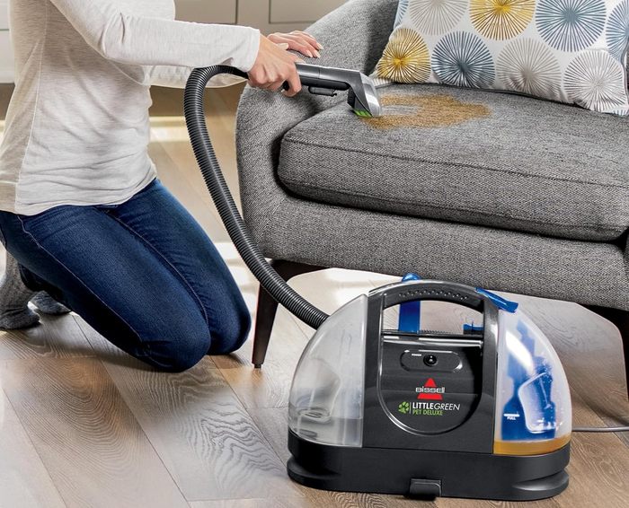 An individual uses a compact vacuum cleaner to eliminate a stain from a sofa. The accompanying text states, 
