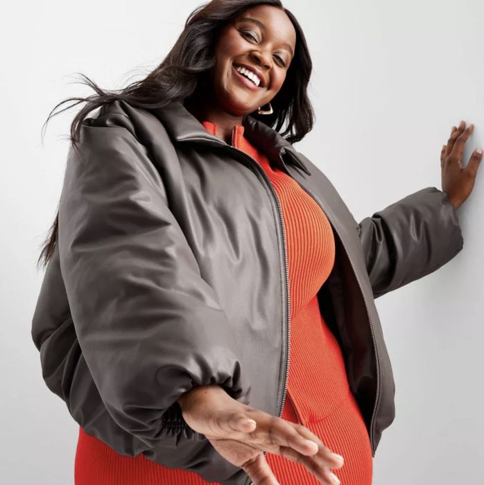 A person wearing a puffy jacket over a ribbed dress, smiling and posing against a simple backdrop
