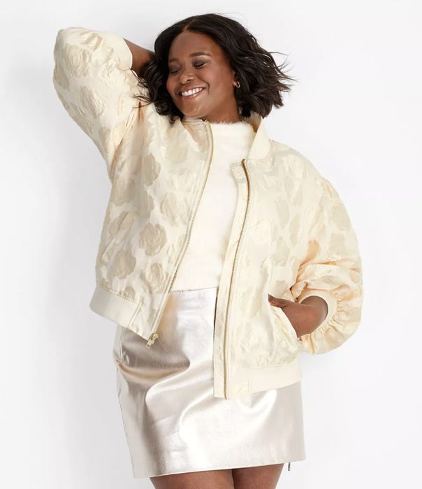 A person smiling while wearing a textured jacket and metallic skirt, posing with one hand behind their head.