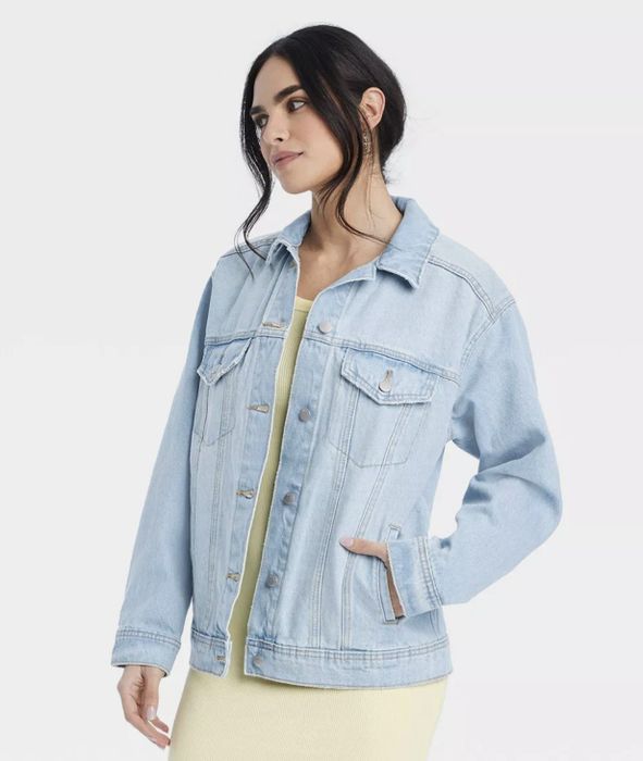A person wearing a denim jacket over a light dress, with hands tucked into the pockets, gazing off to the side.