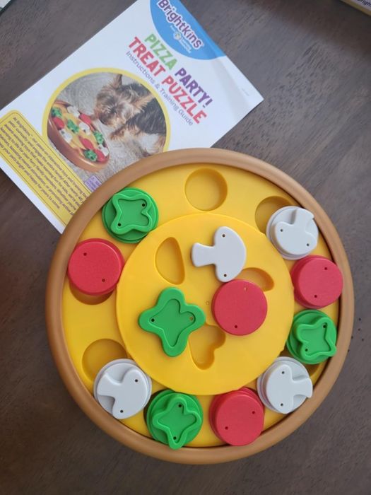 A pizza-themed dog treat puzzle toy with removable pieces, displayed alongside its packaging that includes usage instructions and an image of a dog enjoying treats