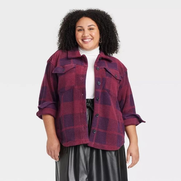 A person beams with a smile, dressed in a purple and red plaid overshirt with sleeves rolled up, paired with a black leather skirt.