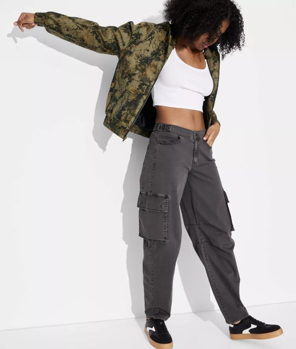 A person in a patterned jacket, white crop top, and dark cargo pants, standing confidently with one arm raised