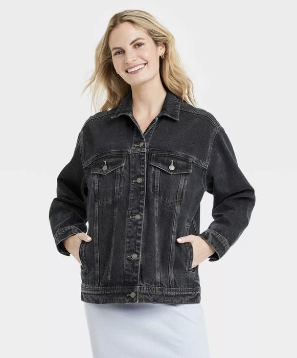 A person smiling while wearing a denim jacket and light skirt, hands in pockets, standing in front of a simple backdrop.