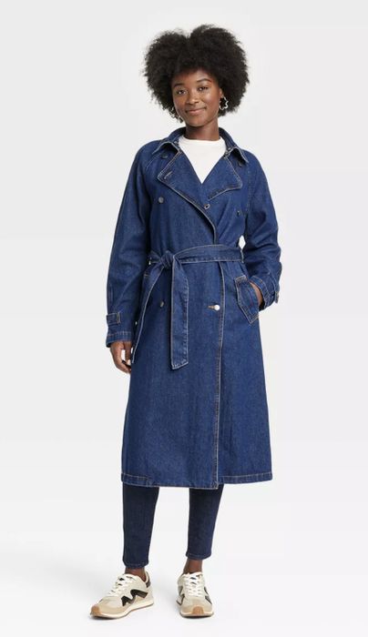 A person wearing a denim trench coat, jeans, and sneakers, standing confidently with a relaxed pose