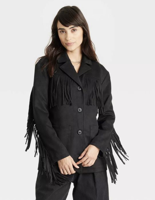 A person in a fringed black jacket over a simple top, posing against a neutral backdrop