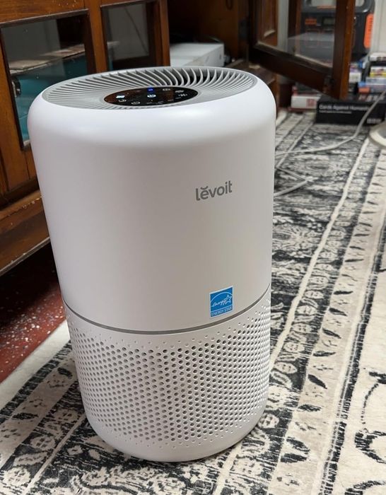 A Levoit air purifier sitting on a patterned rug, surrounded by furniture and other household decor
