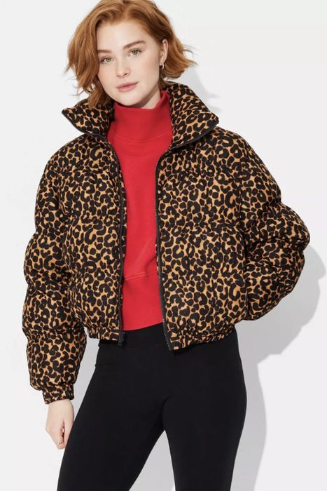 A person wearing a leopard print puffer jacket, red turtleneck, and black pants, striking a pose against a simple backdrop