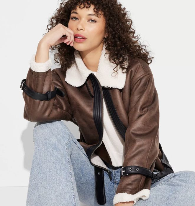 A person sitting casually in a shearling-lined jacket and jeans, their curly hair flowing, with a thoughtful expression