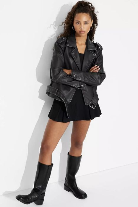 A person dressed in a black leather jacket, black skirt, and knee-high boots, standing confidently against a simple background while gazing at the camera
