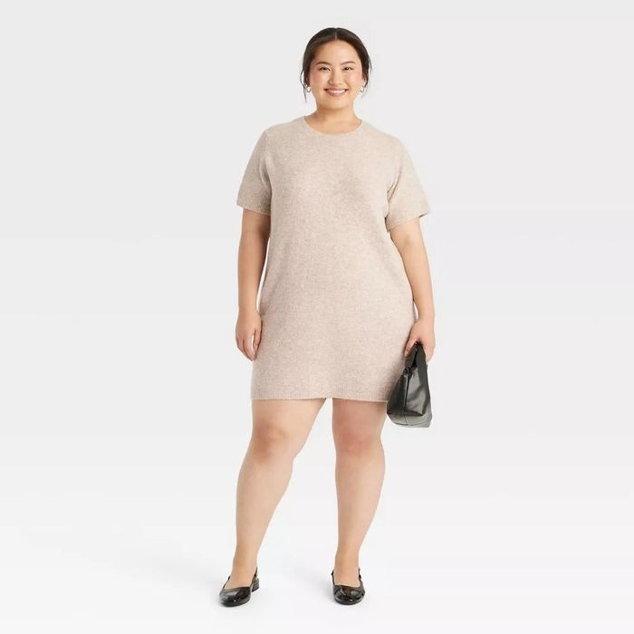A model is seen in a casual, knee-length, short-sleeve dress, holding a clutch bag. She smiles warmly, standing against a neutral backdrop.
