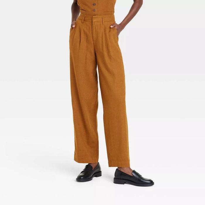A model showcases brown high-waisted trousers featuring front pleats, paired with sleek black loafers, creating a chic look perfect for a shopping feature.
