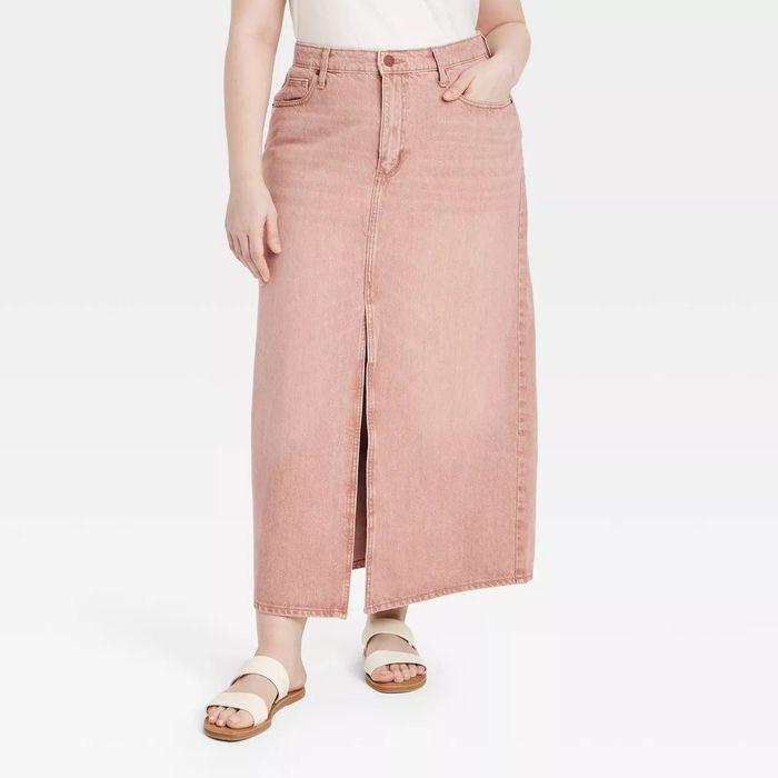 A person (head not visible) is dressed in a long, straight-cut denim skirt featuring a front slit, paired with casual sandals and one hand in their pocket