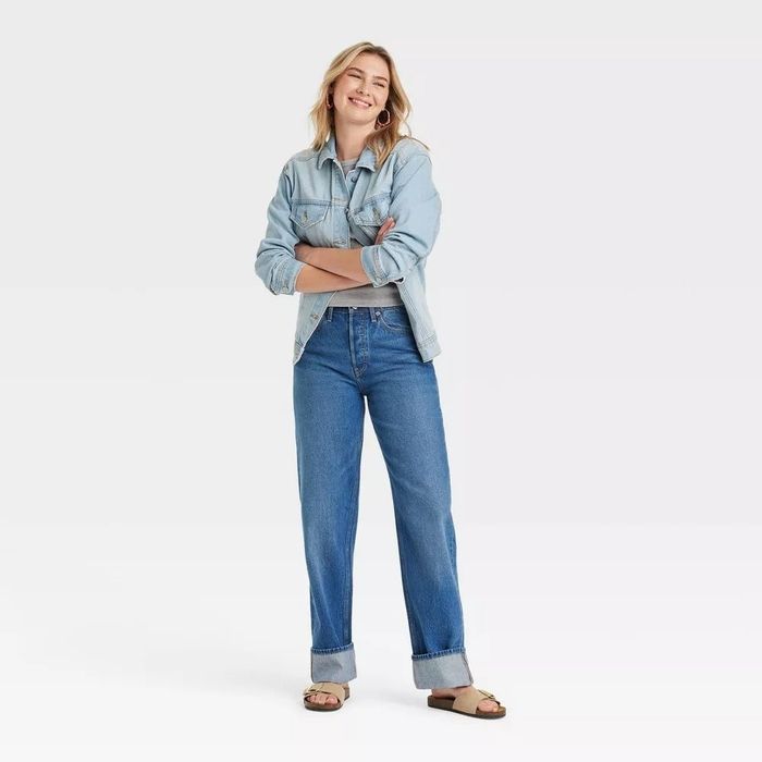 A smiling model stands confidently with arms folded, dressed in a denim shirt, rolled-up jeans, and casual sandals.