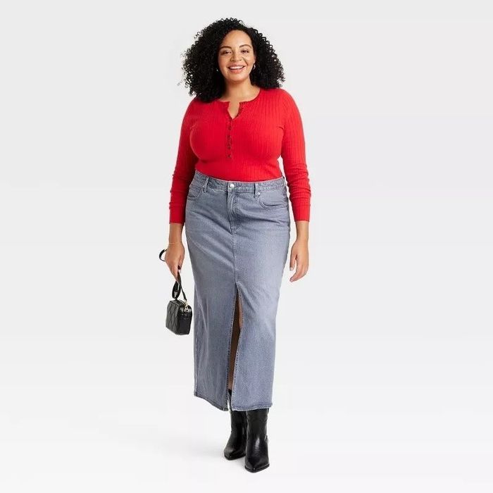 A smiling person wearing a long-sleeve button-up shirt and a front-slit denim skirt, holding a small handbag