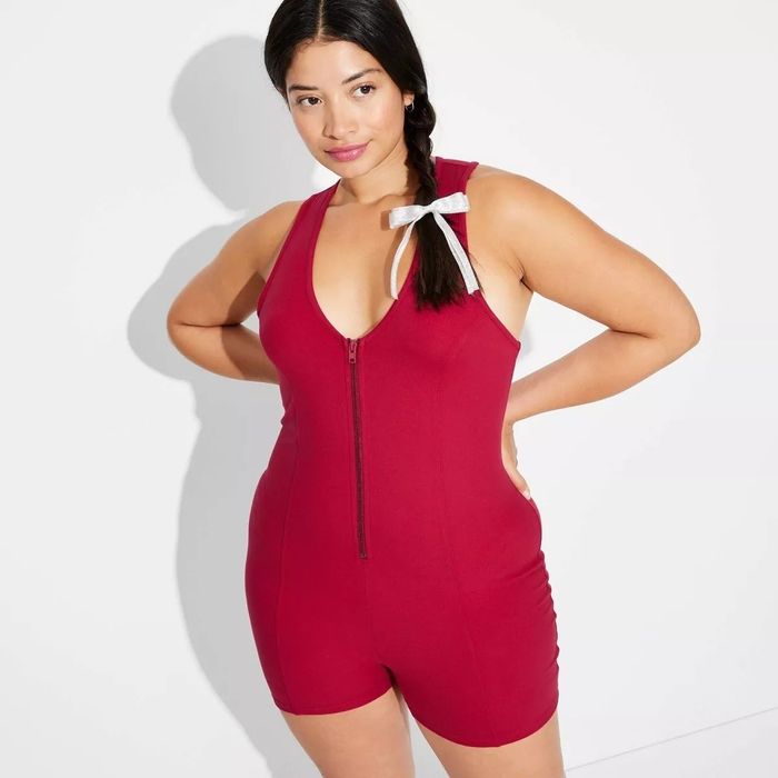 A person strikes a confident pose in a red sleeveless zip-up romper, hands on hips, with a braid tied with a white ribbon.