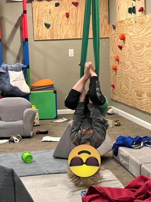 A child is seen hanging upside down from an indoor swing, with a climbing wall visible in the background and toys spread across the floor.