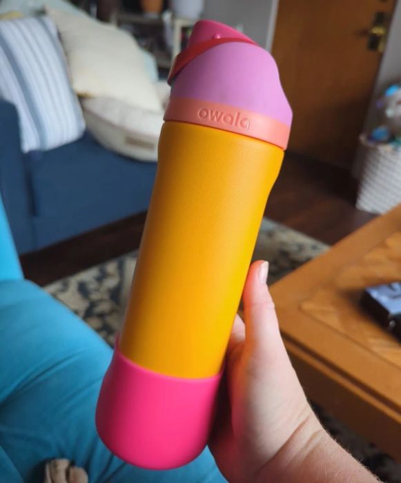 A hand holding a brightly colored Owala water bottle with a pink and orange design, set in a cozy living room.
