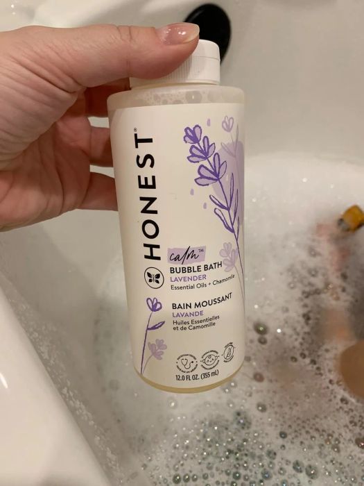 A person holds a bottle of Honest Calm Bubble Bath with lavender and chamomile while enjoying a bubble-filled bath in a tub.