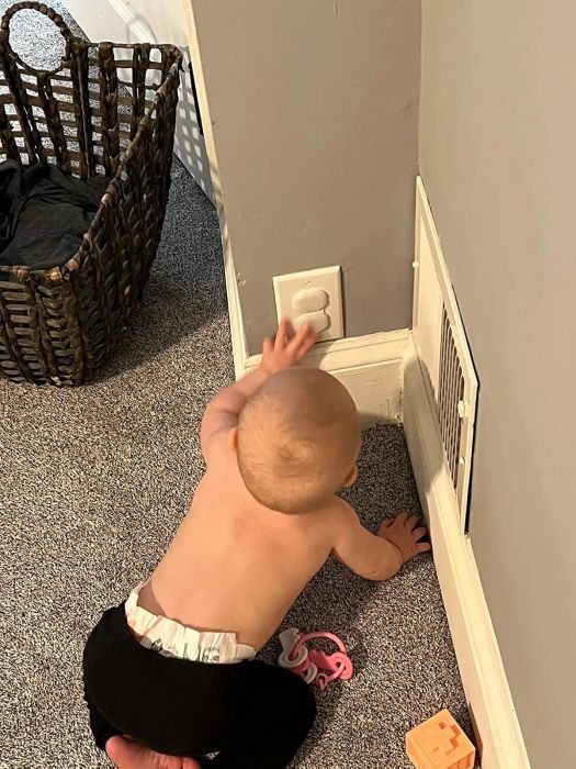 A baby stretches toward an electrical outlet on a carpeted floor, with a wicker basket and a toy nearby.
