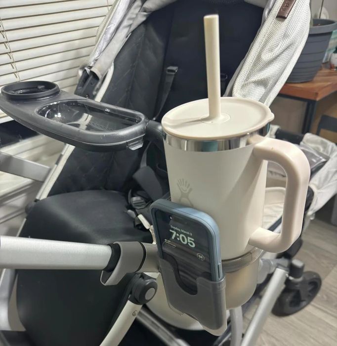 Stroller equipped with a cup holder, featuring a large mug and a phone showing the time as 7:05. Image relevant to shopping for stroller accessories.