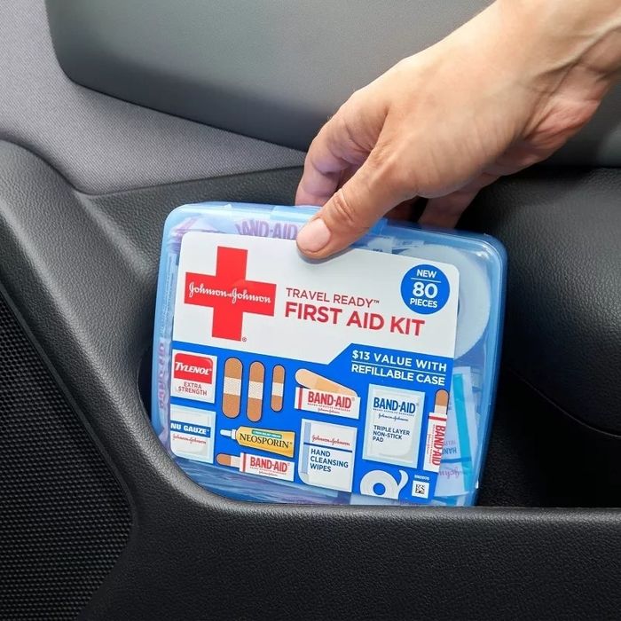 A hand places a Johnson & Johnson travel-ready first aid kit, containing 80 pieces, into the side compartment of a car.