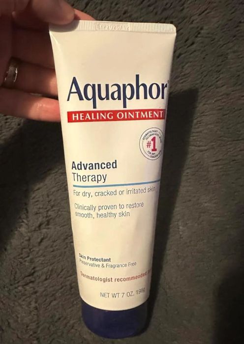 A hand holds a tube of Aquaphor Healing Ointment labeled 'Advanced Therapy,' set against a textured background.