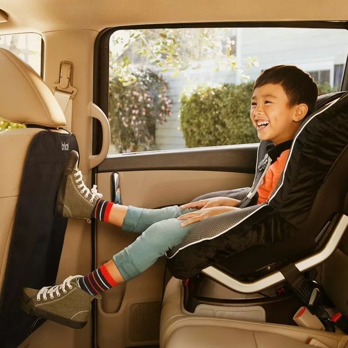 A child smiling in a car seat, dressed in casual clothes and sneakers, with their feet comfortably resting on the seat back, showcasing family travel ease.