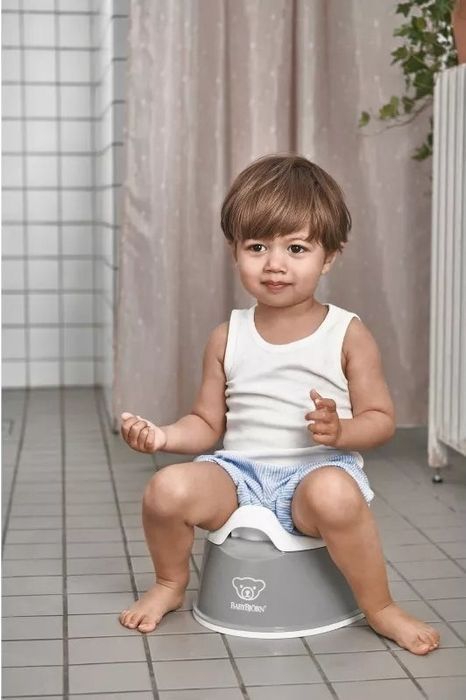 A child seated on a potty in a bathroom, wearing a white tank top and shorts, with a neutral expression on their face.