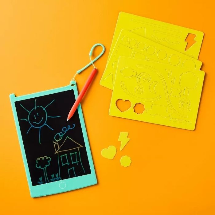 An LCD writing tablet covered in doodles of a sun and house, with yellow stencil sheets displaying various designs, all resting on an orange surface.