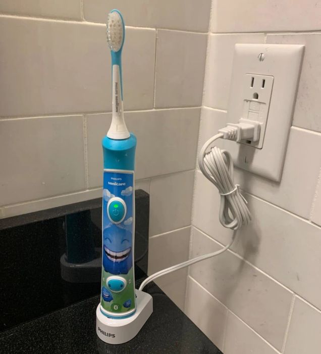 Philips Sonicare kids' electric toothbrush on a charging stand, plugged into a bathroom outlet. The toothbrush has a cheerful smiley face design.