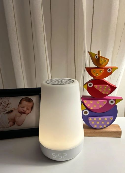 A bedside table featuring a digital alarm clock, a colorful stacked bird toy, and a framed photo of a sleeping baby.