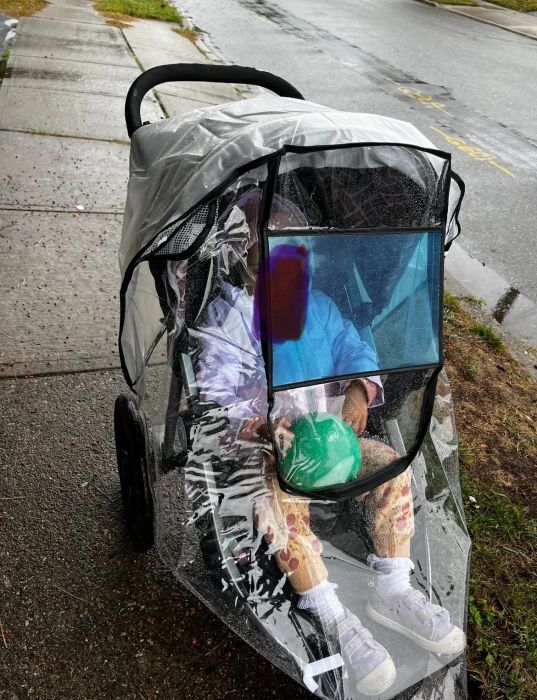 A young child in a stroller with a clear rain cover, holding a green ball. The child wears patterned pants and white shoes, seated on a wet sidewalk.