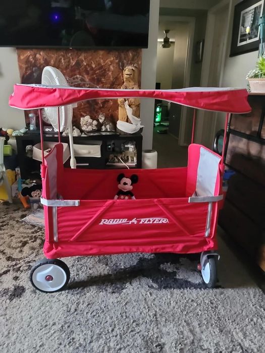 A red Radio Flyer wagon with a canopy is parked indoors, featuring a small Mickey Mouse plush toy seated inside.