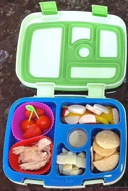 An open bento lunchbox with sections filled with cherry tomatoes, strawberries, cheese cubes, crackers, turkey slices, radish slices, and dip.