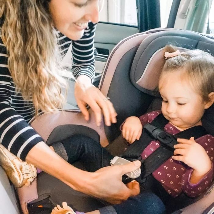 An adult secures a toddler in a car seat. The toddler is dressed in a polka dot shirt and jeans.
