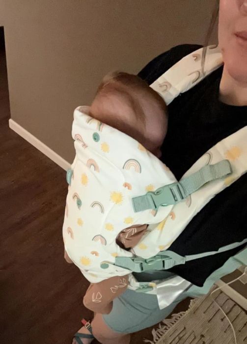 A person carries a baby in a patterned baby carrier, dressed casually in sandals, standing indoors on a wooden floor.