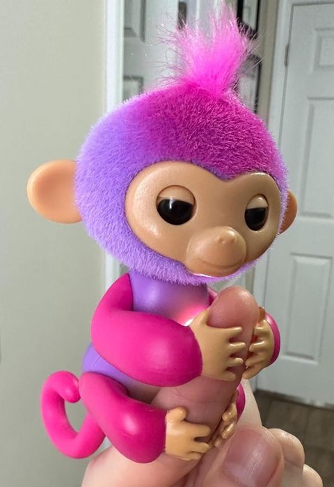 A toy monkey with purple fur, large expressive eyes, and a curled tail, gripping a finger; showcased in a shopping feature