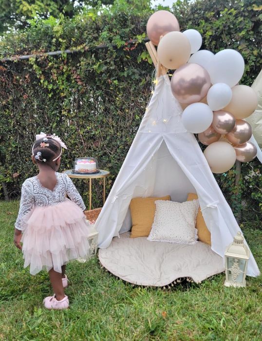 A child wearing a lace top and a pink tulle skirt stands beside a teepee adorned with balloons, next to a cake on a small table in a garden