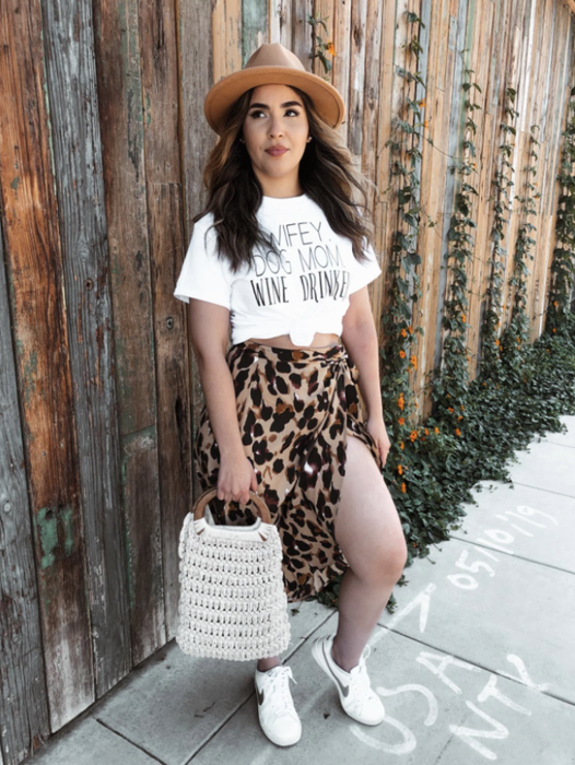 A reviewer is seen wearing the wrap leopard print skirt paired with white sneakers, a graphic T-shirt, and a hat.