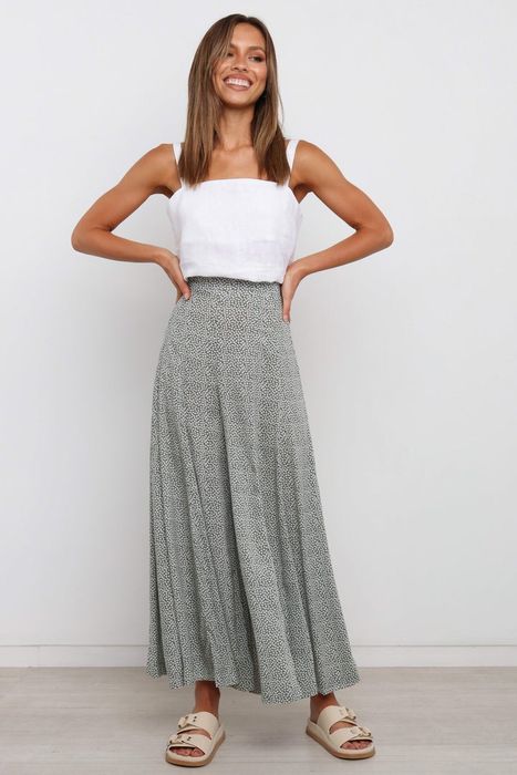 A smiling model in a white top, styled with flowing patterned pants and open-toed sandals.