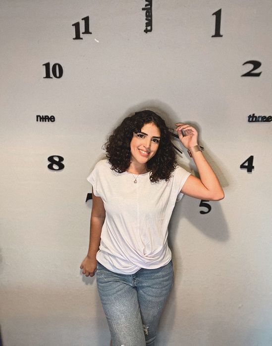 A photo of a reviewer posing in front of a large clock wall, wearing the top paired with jeans.