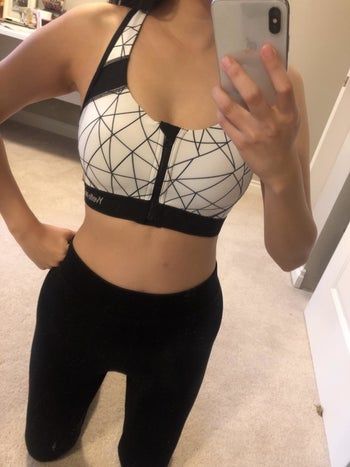 A reviewer taking a selfie in a zip-front black-and-white sports bra and black leggings.