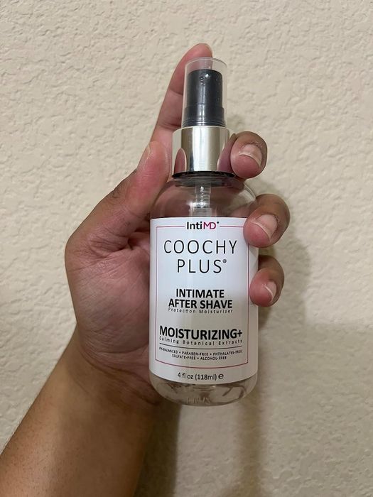 A hand holds the IntiMD Coochy Plus Intimate Moisturizing After Shave, a clear bottle with a white label.