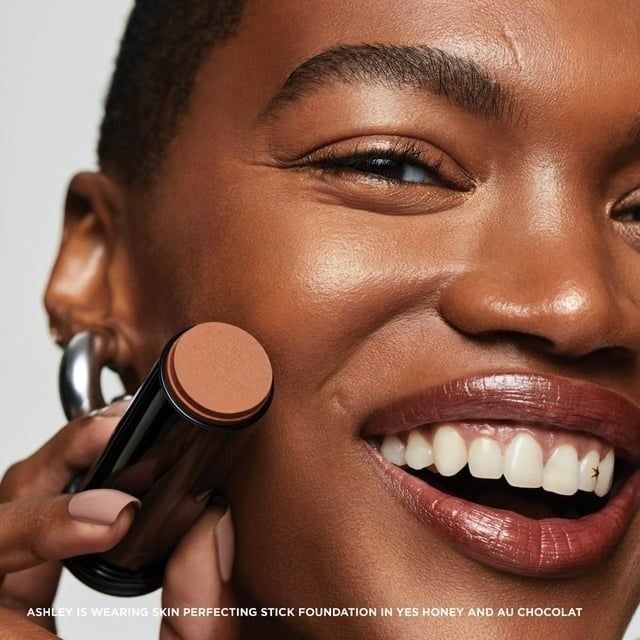 A close-up of a smiling individual holding a foundation stick near their cheek, demonstrating makeup application