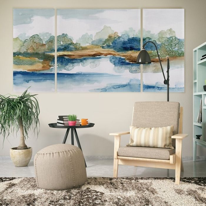 A cozy living room featuring a beige armchair, a pouf, a side table with decorative items, a tall lamp, and a triptych landscape painting adorning the wall.