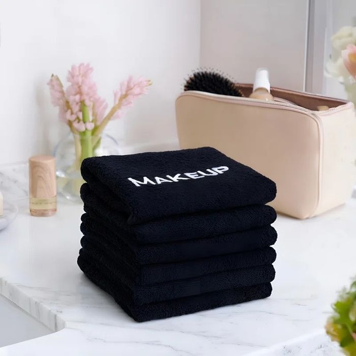 A stack of five black towels, each labeled with the word "MAKEUP", arranged neatly on a marble countertop accompanied by beauty products and fresh flowers.