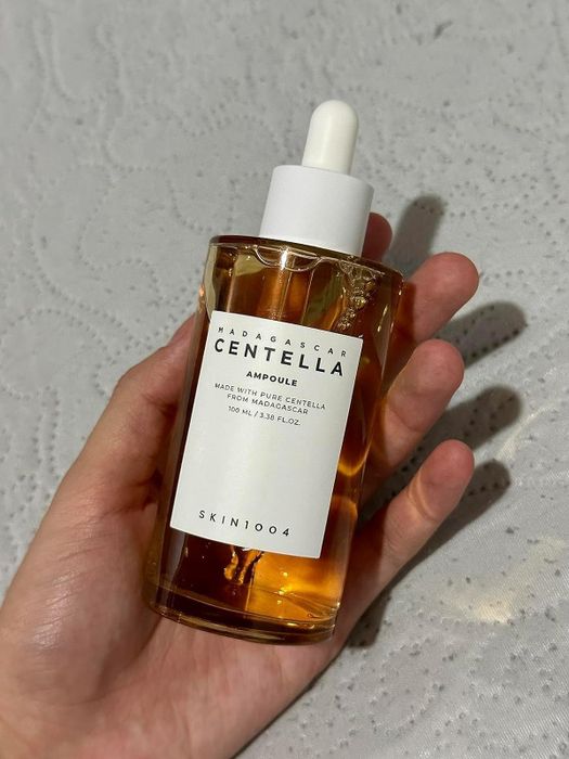 A hand holding a bottle of SKIN1004 Madagascar Centella Ampoule, 100 ml, resting on a textured white surface.